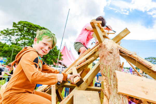 A young boy in an orange onesie with green hair is building with wood.