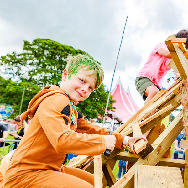 A young boy in an orange onesie with green hair is building with wood.