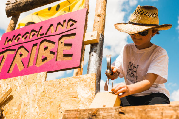 A young boy in a straw hat bangs a nail into some wood with a hammer. There is a pink sign behind him saying Woodland Tribe.