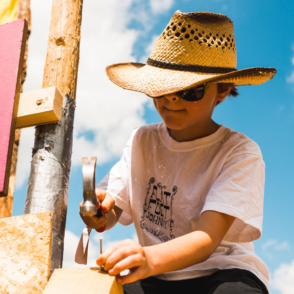 A young boy in a straw hat bangs a nail into some wood with a hammer.