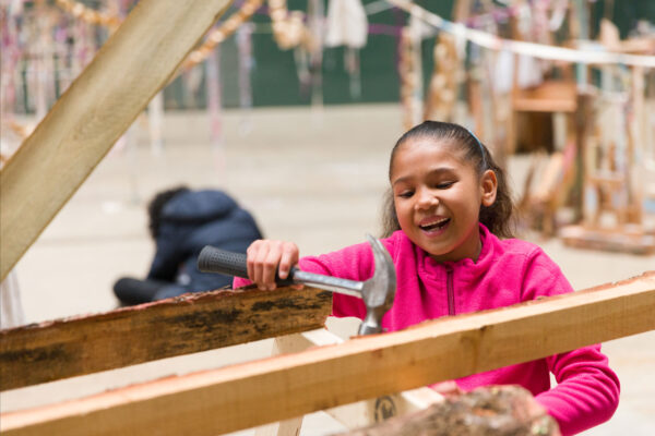 A young girl wearing a bright pink hoodie using a hammer on some wood.