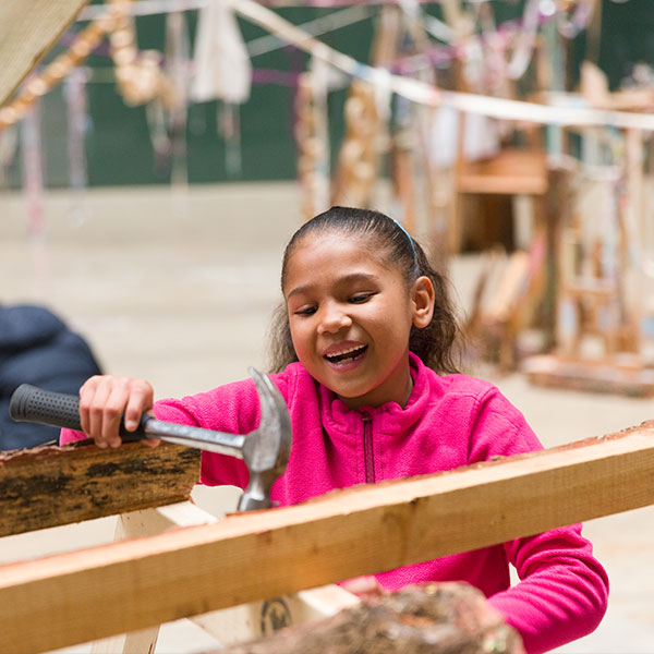 A young girl wearing a bright pink hoodie using a hammer on some wood.