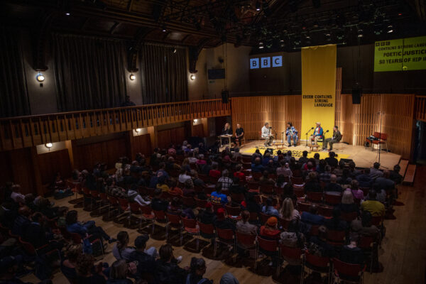 Backs of heads of an audience in a performance hall watching a group of people on stage. The lighting is dark on the audience and light on the stage where there is a yellow banner.