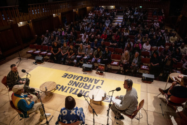 An audience in a performance hall watching a group of people on stage. The lighting is dark on the audience and light on the stage where there is a yellow banner.