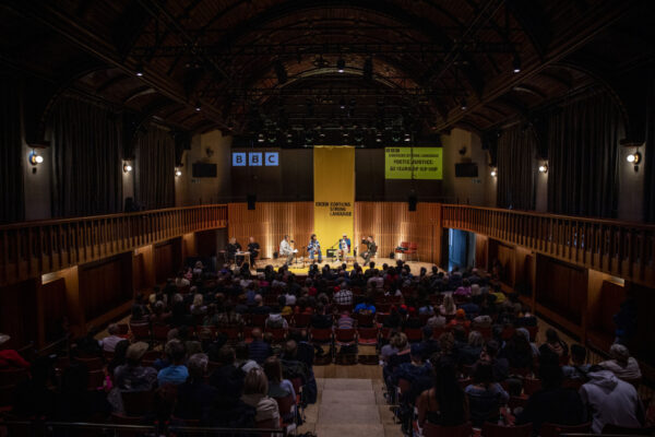 Backs of heads of an audience in a performance hall watching a group of people on stage. The lighting is dark on the audience and light on the stage where there is a yellow banner.