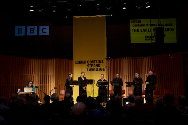 Backs of heads of an audience in a performance hall watching a group of people on stage. The lighting is dark on the audience and light on the stage where there is a yellow banner.