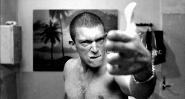 A man looking menacingly into the camera holding his fingers in the shape of a gun. He has a shaved head and is not wearing a top. The picture is black and white.