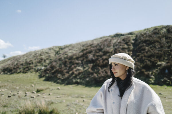 A woman wearing a white coat looks away, with rolling green hills behind her.