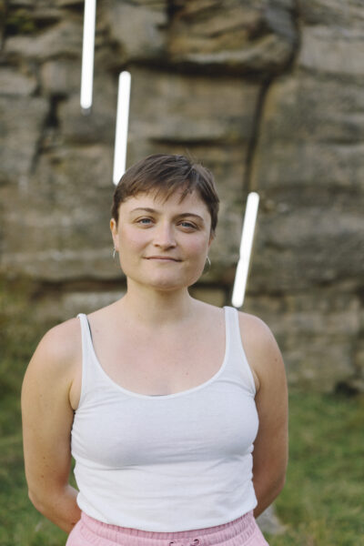 A woman with short brown hair smiles at the camera - she is wearing a white top and there are rocks in the background.
