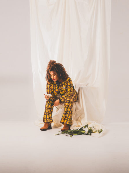 Young lady with curly hair sat down in a yellow patterned suit holding a white flower. The background is white and there is a bouquet of white flowers on the floor next to her feet