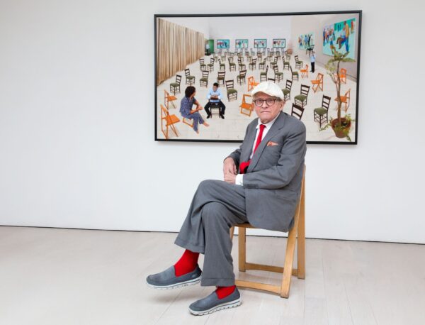 Artist David Hockney sits on a chair in front of one of his paintings. The artwork is hung on a white wall.