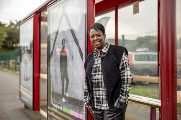 A woman with short black hair, wearing a checked shirt and gilet leans against a picture of herself on a bus stop