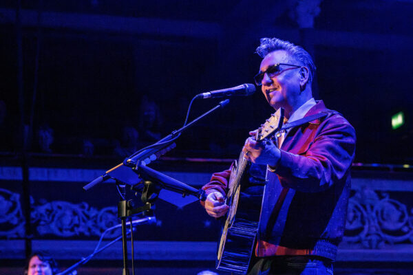 A white man sings into a microphone, he is playing a guitar and wearing a brown jacket