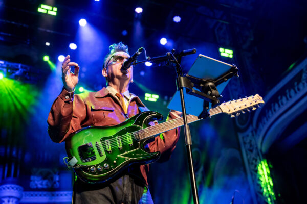A white man sings into a microphone, he is playing a guitar and wearing a brown jacket