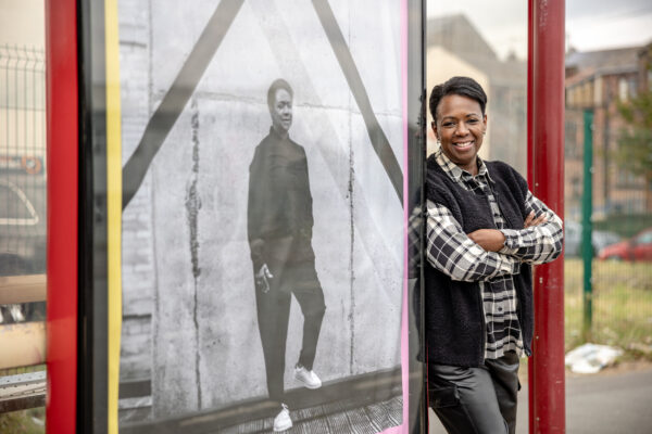 A woman with short black hair, wearing a checked shirt and gilet leans against a picture of herself on a bus stop