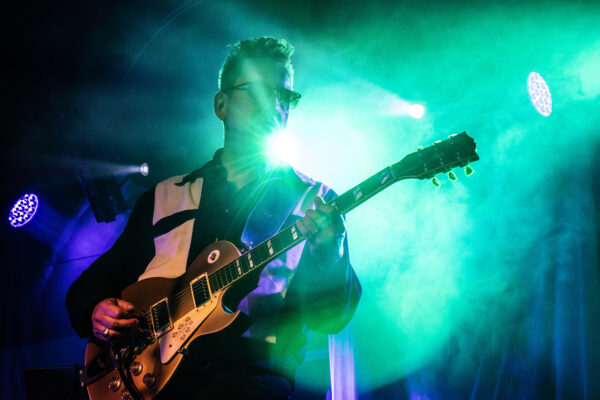 A man playing an electric guitar and wearing sunglasses is backlit by blue and green stage lights.