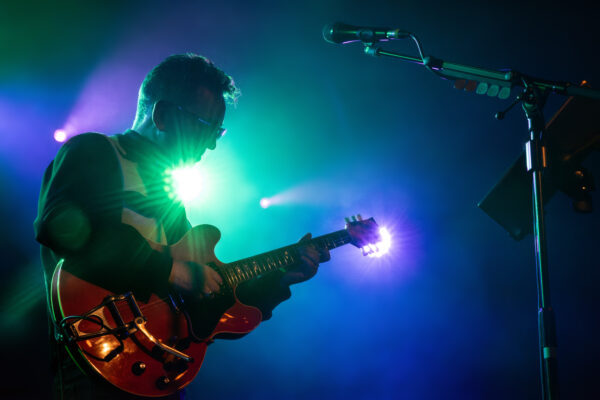 A man playing an electric guitar and wearing sunglasses is backlit by blue and green stage lights.