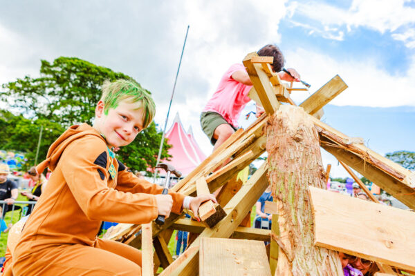 Two young people in bright clothing smile as they climb up a wooden play tower.