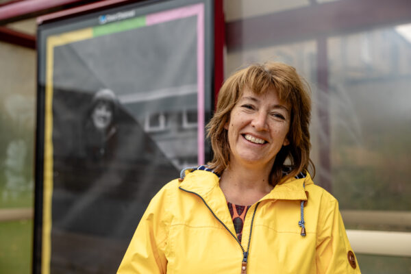 A woman with light brown hair smiles into the camera wearing a bright yellow coat. Behind her is a bus stop with a black and white poster featuring an image of the same woman.