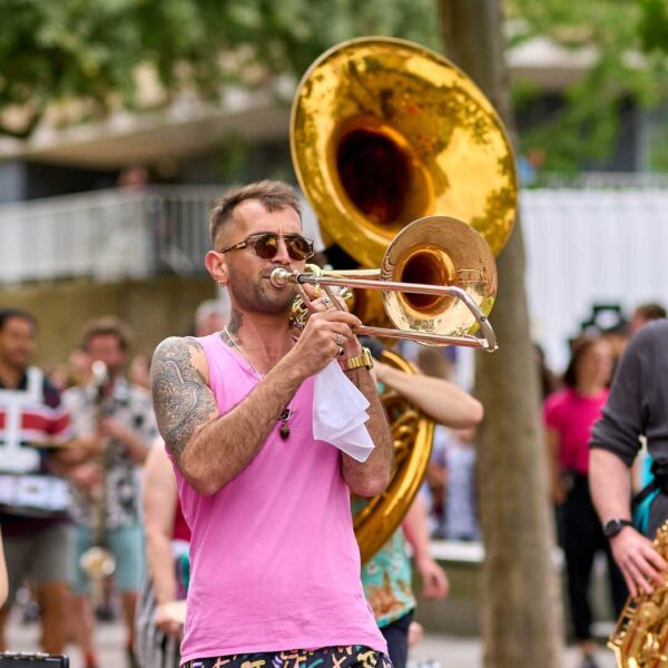 A man walking as part of a parade playing a brass instrument. He is wearing a pink vest and sunglasses.