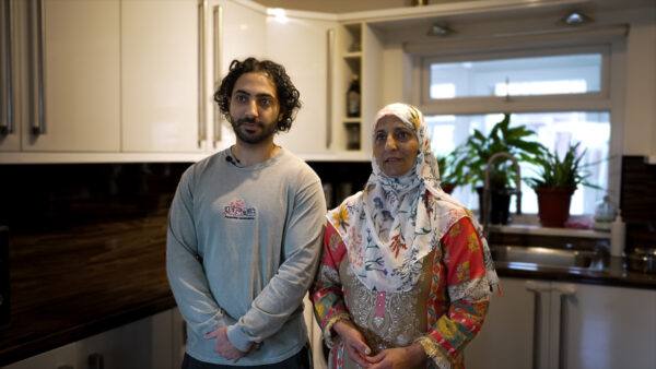 Two people stand in a kitchen - Nabeel and his mum, Nasreen. Nasreen wears a headscarf with an intricate pattern.