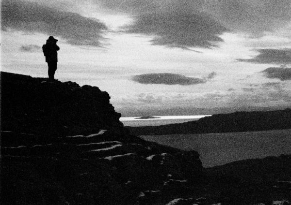 A black-and-white photograph of a person stood on a rocky crag, overlooking the sea at the Isle of Skye.