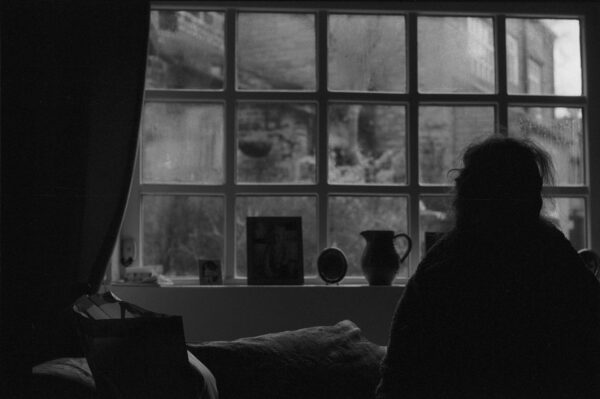 Black-and-white photograph featuring a silhouette of a person sat gazing out of a window. The window sill is decorated with picture frames and a vase.