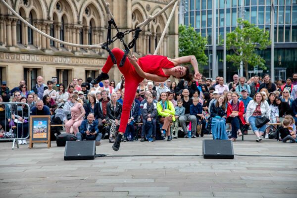 A performer wearing red holds a pose on a rope, suspended above the ground. They are in Bradford City Centre surrounded by a crowd.