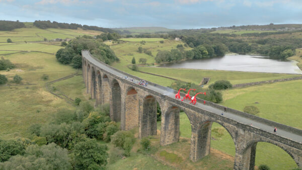 A landscape of a viaduct in the country side with three large red giraffe puppets and a small group walking across the bridge.