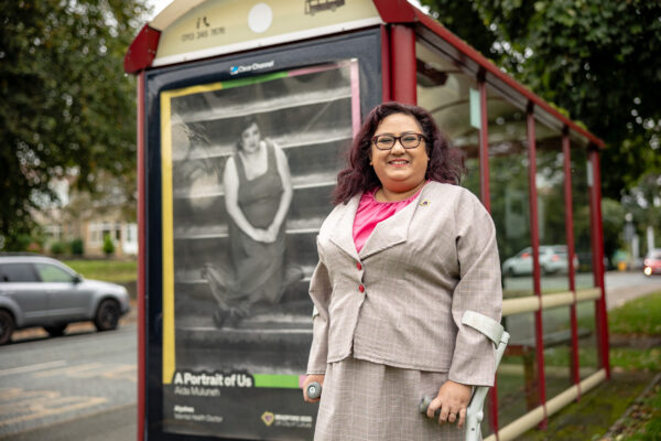 A woman with curly hair and glasses stands next to an image of herself on a bus shelter. She is wearing a pale suit and has crutches.