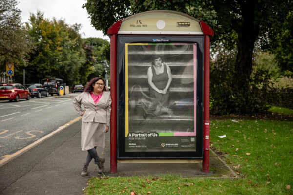 A woman with curly hair and glasses stands next to an image of herself on a bus shelter. She is wearing a pale suit with a pink top.