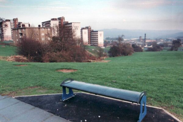 A landscape image of a bench in front of a green field. in the near distance are some high rise flats and in the far distance is the city of Bradford