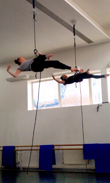 wo people suspended in a bright indoor space, performing aerial movements using harnesses attached to the ceiling. One person floats horizontally with their arms extended, while the other is positioned slightly higher, reaching out in a graceful pose. A window in the background lets in natural light.