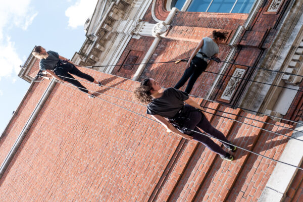 Three people wearing harnesses are suspended on ropes, performing a vertical dance against the side of a red brick building with ornate architectural details. The performers appear confident and focused, with a blue sky in the background.