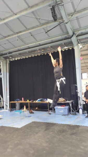 A woman wearing a harness performs mid-air with her arms raised triumphantly in an indoor rehearsal space. The ceiling features exposed beams, and the backdrop includes a black curtain with a table holding various equipment and supplies. The floor is covered in black mats.