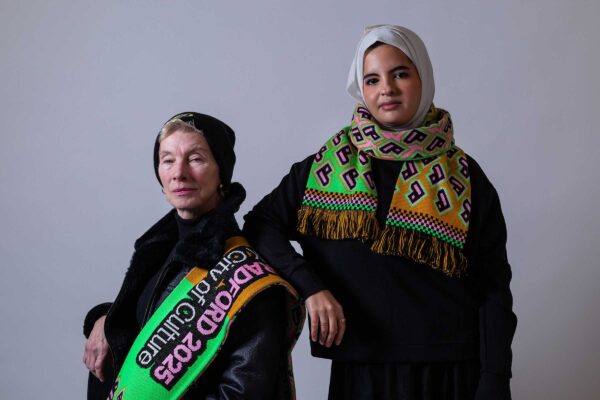 Two models in black clothes wear the colourful Bradford 2025 branded scarf. They stand against a white background. The model on the left is sitting down. The model on the right is standing, and wears a headscarf.