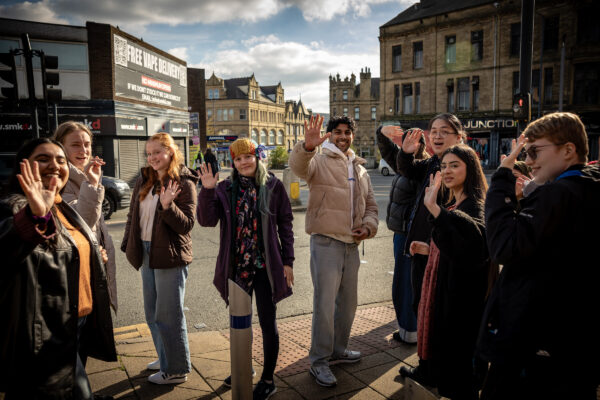Group of young people stood outside in a semi circle waving and smiling to the camera.