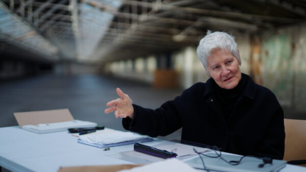 Artist Ann Hamilton, with short white hair and wearing a dark coat, gestures thoughtfully while seated at a table covered in papers, a tablet, and writing tools. She is in the expansive, light-filled interior of Salts Mill in Saltaire, a historic industrial building with high ceilings and exposed beams.
