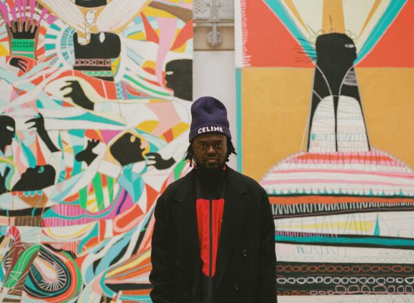 Headshot of man in beanie and black coat infront of his lare pieces of colourful abstract artwork