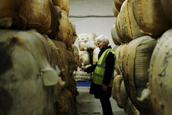 A lady with white hair in a hi-vis jacket stands between two stacks of giant bales of wool