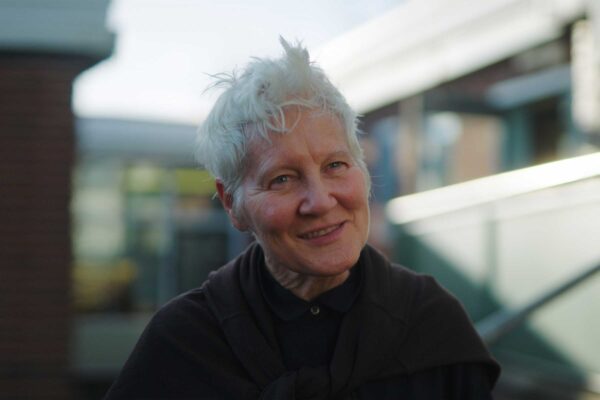 A lady with short grey hair stands outside smiling. Her gaze is to the right of the camera.