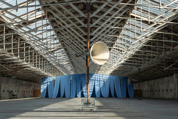 A wide shot of a metal horn on a pole in front of a giant blue curtain. They are inside the roof of an old mill.