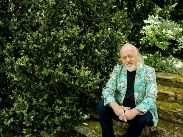 Bill Bailey sits in front of a large bush. He wears an aquamarine jacket.