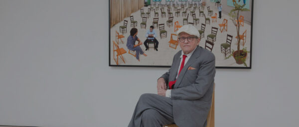 Artist David Hockney sits on a chair in a gallery, in front of one of his paintings.