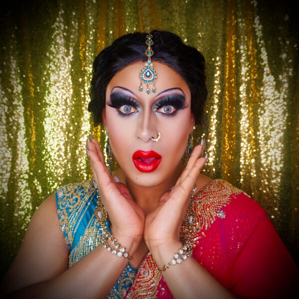 A headshot of a drag queen wearing traditional red and blue south asian clothing and a blue jewellery head piece.