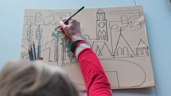 Hand colouring in, in green papers, a line drawing of buildings on a cardboard piece of paper