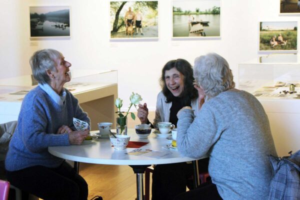 Three older women sit around a table in an art gallery. They are laughing and drinking tea.