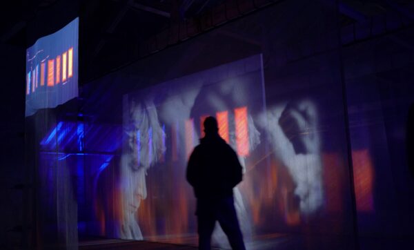 The black form of a figure interrupts projected faces on pieces of fabric hanging from the ceiling.