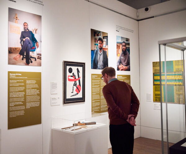 Man in an exhibition looking at a calligraphy pen in a glass case. On the wall is three images of men with bios underneath.