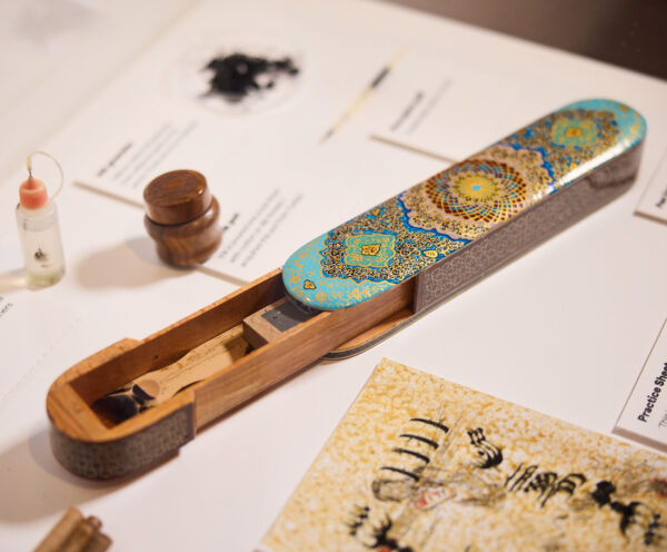 Decorative calligraphy pen case with a sliding wooden lid, and pen inside. Surrounding is an ink bottle, wooden inkwell and a piece of paper with hand written calligraphy on it.